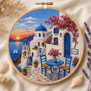 Può includere: Un telaio da ricamo circolare contenente una scena a punto croce di un villaggio su un'isola greca al tramonto. Il disegno presenta edifici bianchi con cupole blu, un tavolo e sedie blu, fiori rosa e un cartello con la parola greca Agapi.