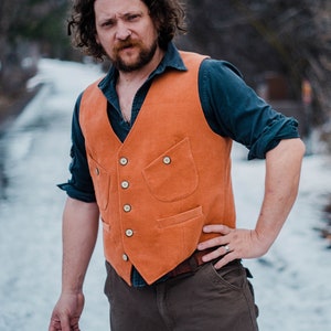 Puede incluir: Un hombre con un chaleco marrón sobre una camisa azul, pantalones marrones y un anillo de plata en la mano izquierda. Está de pie en un área nevada con árboles en el fondo.