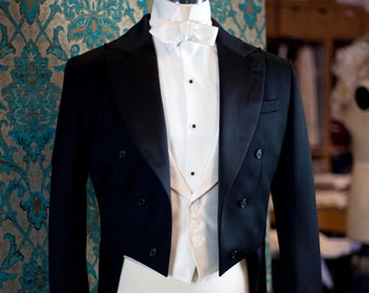 Puede incluir: Un esmoquin negro formal con camisa blanca, chaleco y pajarita, exhibido en un maniquí. La chaqueta tiene un diseño cruzado con botones negros y corte de frac. El fondo presenta un papel pintado estampado.