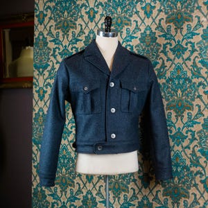 May include: A dark blue jacket with a tailored fit, displayed on a mannequin. The jacket features a notched collar, multiple pockets, and button closures. The background is a patterned wallpaper with teal and gold floral designs.
