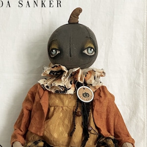 Puede incluir: Una muñeca artesanal de arte popular con cabeza de calabaza, vestida con un atuendo marrón y naranja con polainas a rayas blancas y negras. La muñeca está sentada en un carrete de hilo y tiene un cuello con estampado blanco y negro.