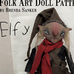 May include: Folk art doll pattern for an elf doll with a red scarf, brown pants, and a brown hat. The doll is sitting on a white wooden box. The text "Folk Art Doll Pattern" and "By Brenda Sanker" is written on the white background. The text "Elfy" is written in black marker on the white background.