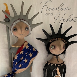 May include: Two folk art dolls, one dressed as the Statue of Liberty with a red, white, and blue scarf and the other dressed in black with a witch tag. The dolls are both standing on a white fabric background. The text "Freedom and Hekate" is written in cursive above the dolls.