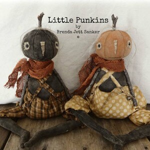 May include: Two handmade rag dolls dressed as pumpkins. The dolls have black bodies, orange pumpkin heads, and are wearing plaid and polka dot fabric. The dolls are sitting on a wooden surface. The text "Little Punkins by Brenda Jett Sanker" is above the dolls.