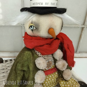 May include: A handmade snowman doll wearing a green jacket, red scarf, and white pom-poms. The snowman has a black top hat with the words "Keeper of the Snow" written on it. The snowman is sitting on a wicker chair.