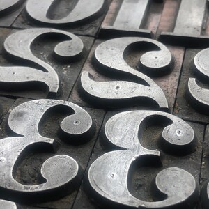 May include: Close-up of a collection of metal letterpress printing blocks, each featuring a different letter of the alphabet. The letters are arranged in a random order, creating a visually interesting pattern.