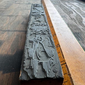 May include: A long, rectangular metal letterpress printing block featuring a raised, intricate relief design of a figure in a flowing garment. A wooden ruler placed alongside shows the block measures approximately 22 inches in length.