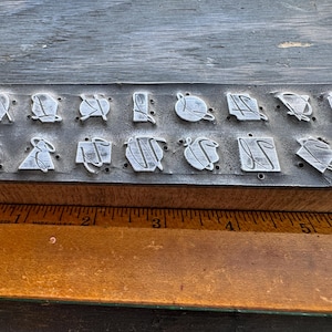 May include: A rectangular rubber stamp block featuring sixteen distinct raised metal icons arranged in two rows. The block is positioned against a wooden ruler, showing a length of approximately 5.5 inches. Each icon displays a unique geometric shape.