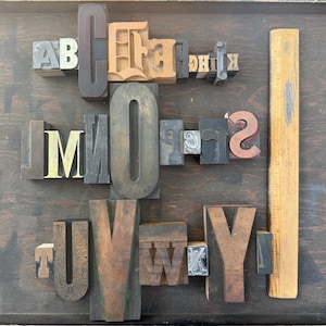 May include: A collection of vintage wooden letterpress printing blocks, including letters A through Y, numbers, and symbols. The blocks are various sizes and colors, with a wooden ruler on the right side. The letters are arranged on a dark wooden surface.