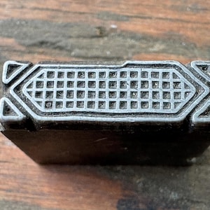 May include: A rectangular metal printing block featuring a raised grid pattern on its top surface. The design is enclosed within a geometric border with triangular accents at each corner. The metal has a dark, weathered finish with silver highlights.