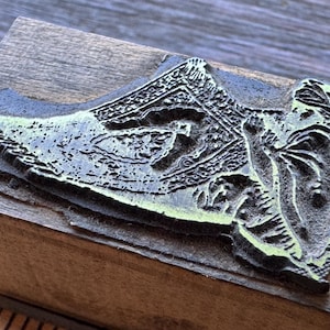 May include: A vintage wood block stamp with a detailed relief image of a person holding a shield and a patterned object. The stamp's relief is a dark gray color with hints of green, mounted on a rectangular wooden block.