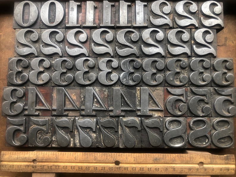 May include: A collection of vintage metal letterpress printing blocks featuring the numbers 0 through 9. The blocks are arranged in rows and are made of a dark gray metal with a worn, textured surface.