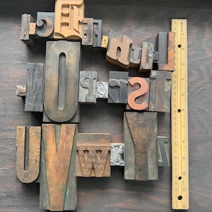 May include: A collection of various wooden and metal letterpress printing blocks featuring assorted uppercase letters, arranged in a grid-like formation next to a wooden ruler that measures up to 12 inches in length, showing the scale of the blocks.