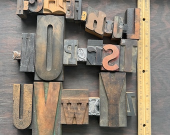 May include: A collection of various wooden and metal letterpress printing blocks featuring assorted uppercase letters, arranged in a grid-like formation next to a wooden ruler that measures up to 12 inches in length, showing the scale of the blocks.