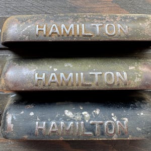 May include: Three vintage metal drawer pulls with a weathered finish, each featuring the embossed text HAMILTON on the front. These rectangular bin-style handles have mounting holes on each side and show signs of oxidation and paint wear.