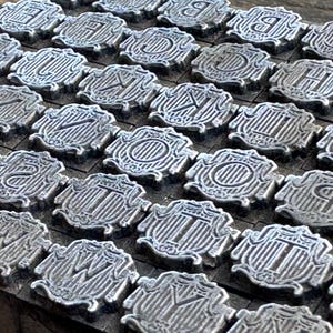May include: Close-up of a collection of silver letterpress printing blocks. Each block features a different letter of the alphabet, set within a decorative shield-like frame. The blocks are arranged in neat rows.