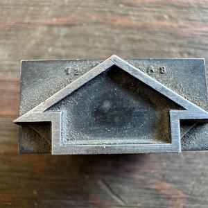 May include: A rectangular metal stamp with a house-shaped design. The stamp has a dark, aged appearance with the numbers "12" and "AB" etched into the top. The house outline is a lighter color, creating a contrast.