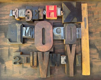 Può includere: Una collezione di blocchi tipografici vintage in legno e metallo con varie lettere maiuscole, tra cui una grande H rossa e una prominente O marrone. Un righello in legno da 30 cm è posizionato verticalmente sul lato destro.