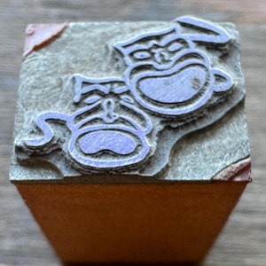 May include: A square rubber stamp featuring a raised design of two theatrical masks, one smiling and one frowning, set against a textured gray background. The stamp is mounted on a solid, light brown wooden block base with visible corner wear.