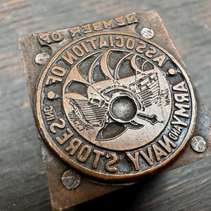 May include: A vintage brass stamp with the text "MEMBER OF ASSOCIATION OF ARMY AND NAVY STORES INC." and a graphic of a ship's wheel and anchor.