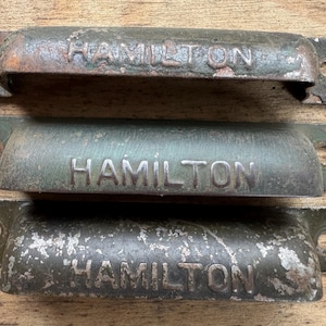 Puede incluir: Tres tiradores de cajón de metal vintage con el nombre "HAMILTON" grabado en relieve. Los tiradores tienen un aspecto desgastado y envejecido con tonos verdes, marrones y plateados. Cada tirador tiene dos orificios de montaje.