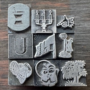 May include: A collection of nine metal letterpress printing blocks arranged in a grid. The blocks feature embossed designs including a mortar and pestle, a candelabra, a pointing hand, the letter U, a flag with the number 240, a telephone, a turkey, a cat, and a tree.
