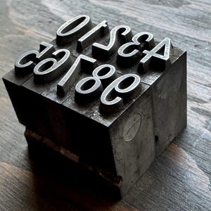 May include: A rectangular metal letterpress block featuring raised characters. The top surface displays the letters O, I, S, E, A, C, G, and the number 8 in a serif typeface. The block has a dark, weathered metallic finish with visible signs of use.