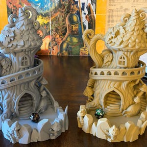 May include: Two fantasy-themed dice towers, one gray and one tan, resembling whimsical treehouses. Each tower features a detailed design with arched doorways, mushroom accents, and a circular balcony. Dice are visible in the base of each tower.