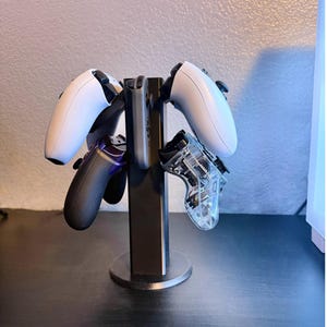 May include: A black stand with four video game controllers. Two white controllers are on the left and right sides of the stand. A black controller with purple accents is on the left side of the stand. A clear plastic controller is on the right side of the stand.