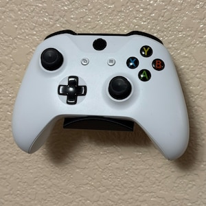 May include: A white Xbox controller with black buttons and directional pad. The controller has colored buttons labeled A, B, X, and Y. The controller is mounted on a black stand.