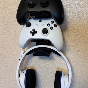 May include: A black and a white Xbox controller are mounted on a wall, with a white headset below. The controllers have colored buttons and black joysticks. The headset has white earcups with black and blue accents.
