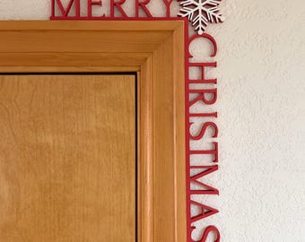 May include: A red wooden Christmas decoration with the words "MERRY CHRISTMAS" and a white snowflake. The decoration is placed on the corner of a wooden door frame and a white wall.