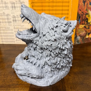 May include: A gray wolf head sculpture with an open mouth, revealing sharp teeth. The sculpture has detailed fur texture and is likely a decorative item or a container for small objects. The wolf is set on a rocky base.
