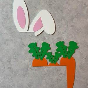 May include: White bunny ears with pink inner ears and a set of orange carrots with green tops. The Easter-themed decorations are made of wood and are set against a gray background.