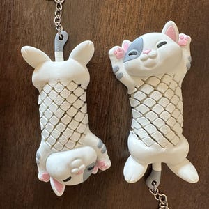 May include: Two white cat and bunny keychains with silver key rings. The cat has gray and pink accents, while the bunny has gray and pink accents. Both have a textured, scale-like design on their bodies.