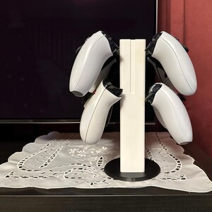 May include: White plastic stand with four slots for holding video game controllers. The stand has a black base and is designed to hold controllers upright.