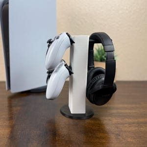 May include: A white gaming console stand with a black base, holding two white game controllers and a pair of black headphones. The stand is on a dark wooden surface, with a white gaming console in the background.