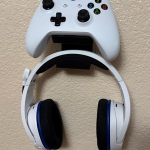May include: White Xbox controller and headset mounted on a wall. The controller has black buttons and a directional pad. The headset is white with black and blue accents.