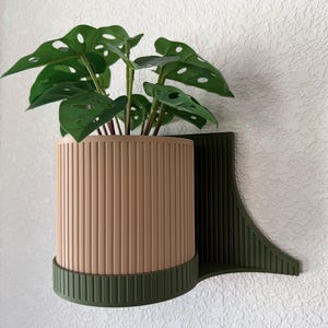 May include: A decorative wall-mounted planter with a tan, vertically ribbed cylindrical pot and a dark green bracket. The planter holds a green plant with large, uniquely shaped leaves. The design is modern and minimalist.