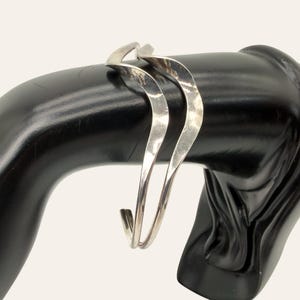 May include: A silver-toned cuff bracelet featuring a double-layered, twisted, and undulating wave design. The metal surface has a polished, reflective finish that catches the light, creating a fluid, continuous shape that wraps around the wrist.