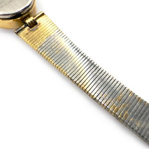 May include: Close-up of a vintage wristwatch with a gold-toned case and a silver and gold-toned flexible band. The watch face is round with a white dial. The band is made of small, rectangular metal links.