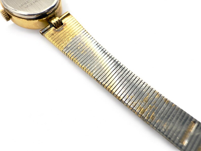 May include: Close-up of a vintage wristwatch with a gold-toned case and a silver and gold-toned flexible band. The watch face is round with a white dial. The band is made of small, rectangular metal links.
