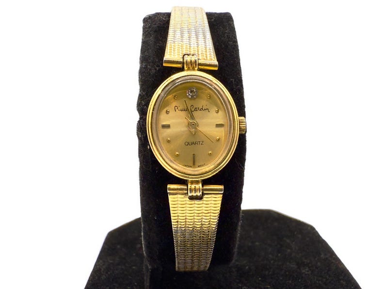 May include: Gold-toned Pierre Cardin quartz watch with a textured band. The watch features an oval face with a gold dial, a small diamond accent, and the brand name. The band has a woven design.