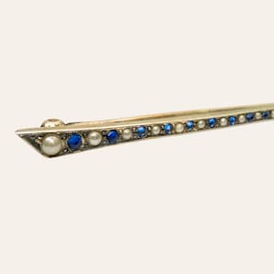 Puede incluir: Un broche de barra largo y delgado hecho de metal, que presenta un patrón repetitivo de pequeñas perlas blancas redondas y piedras preciosas de color azul vibrante dispuestas en una fila lineal a lo largo de la pieza, con un extremo puntiagudo.