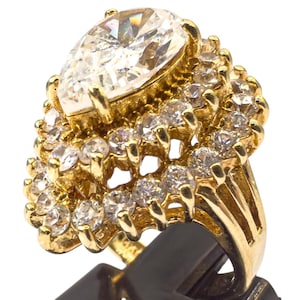Puede incluir: Un anillo dorado con una gran piedra central en forma de pera, rodeada de numerosas piedras claras más pequeñas. El diseño del anillo es multicapa. Las piedras brillan sobre un fondo blanco.