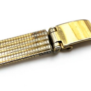 May include: Close-up of a gold-toned watch band with a folding clasp. The band is made of small, rectangular links, and the clasp is rectangular with a brushed finish. The band is on a white background.