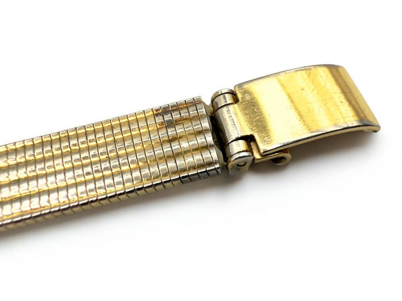 May include: Close-up of a gold-toned watch band with a folding clasp. The band is made of small, rectangular links, and the clasp is rectangular with a brushed finish. The band is on a white background.