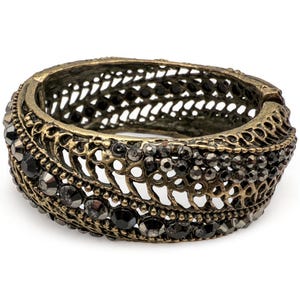 Puede incluir: Un brazalete articulado de tono dorado antiguo con intrincados calados y gemas negras incrustadas. El brazalete presenta un diseño de filigrana detallado y se encuentra sobre un fondo blanco, mostrando su estilo ornamentado.