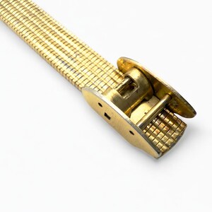 May include: A gold-toned metal watch band with a mesh-like design. The band features a clasp with a rectangular grid pattern and two oval-shaped end pieces. The band is set against a white background.