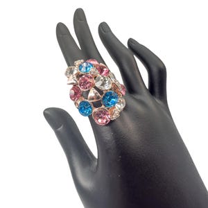 Peut inclure: Une bague imposante avec un groupe de pierres précieuses colorées. La bague a un anneau doré et est ornée de cristaux roses, bleus et transparents. La bague est présentée sur une main noire, mettant en valeur sa taille et son design.